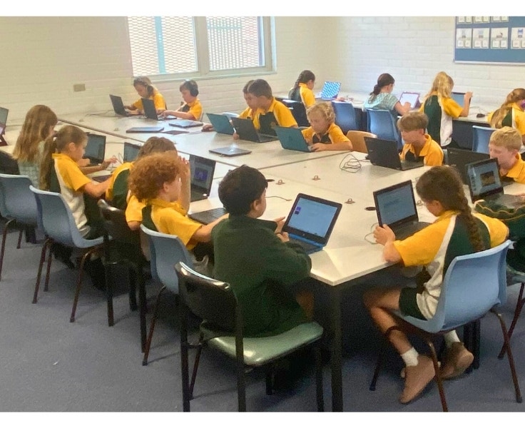 Students wearing green and gold uniform using laptops while sitting around a large group of tables