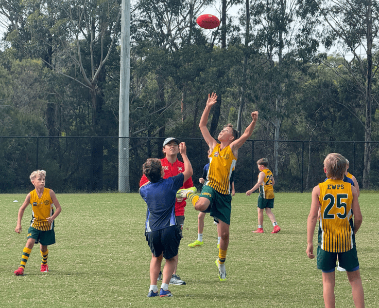 Student in green and gold AFL uniform jumping up in mid air to reach an AFL ball while other team mates watch