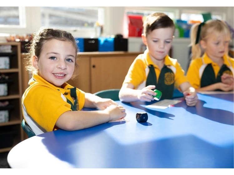 A girl wearing gold and green polo looking at the camera while another boy and girl play with colourful dice on a blue table