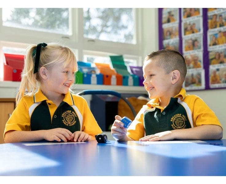 Girl and boy wearing green and gold school uniform sitting at a blue desk playing with coloured dice