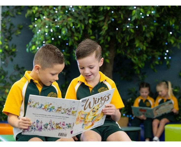 Two boys wearing green and gold school uniform sitting in the library reading a book with girls sitting in distance
