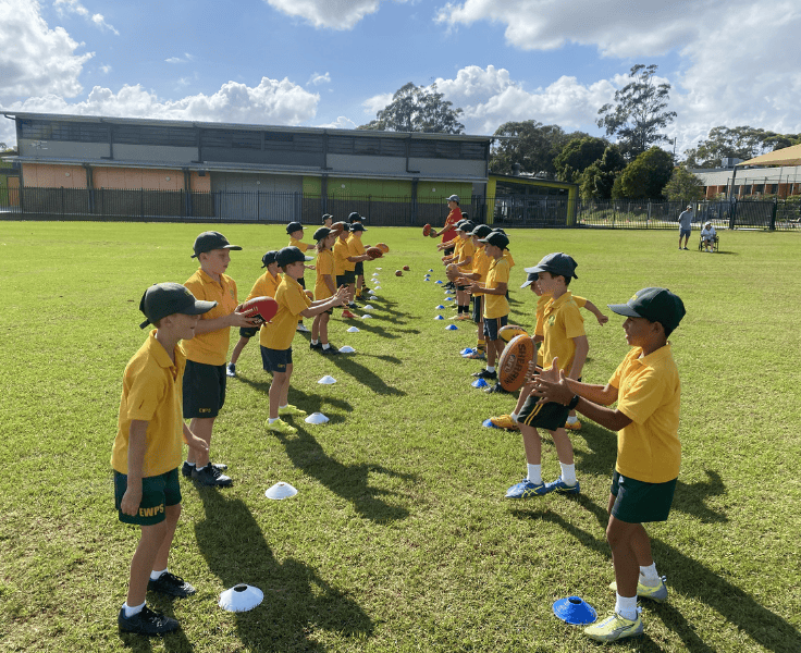 Students wearing gold shirts, green shorts and green caps facing each other in two lines passing footballs to each other on a grassy oval