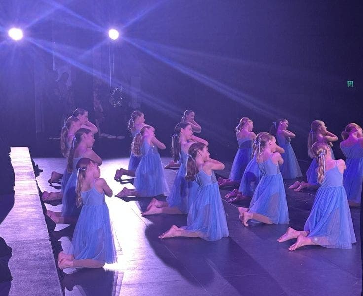 Female dancers in blue dresses kneeling on stage with lights shining on them