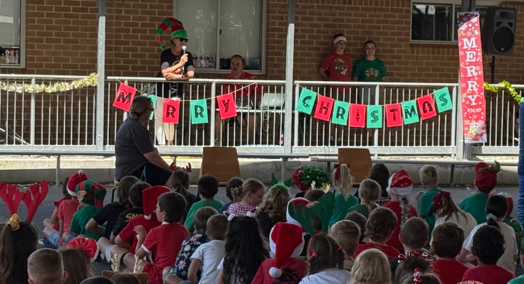Students dressed in Christmas outfits seated in ground facing teacher standing on verandah speaking into microphone with merry Christmas signage below