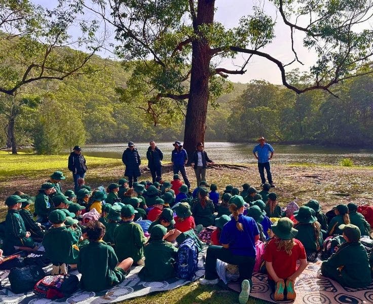 A large group of students sitting on grassed area in front of river surrounded by trees