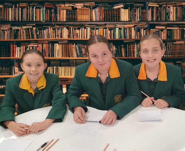 Three female students wearing green blazers sitting at a desk in front of a wall of books